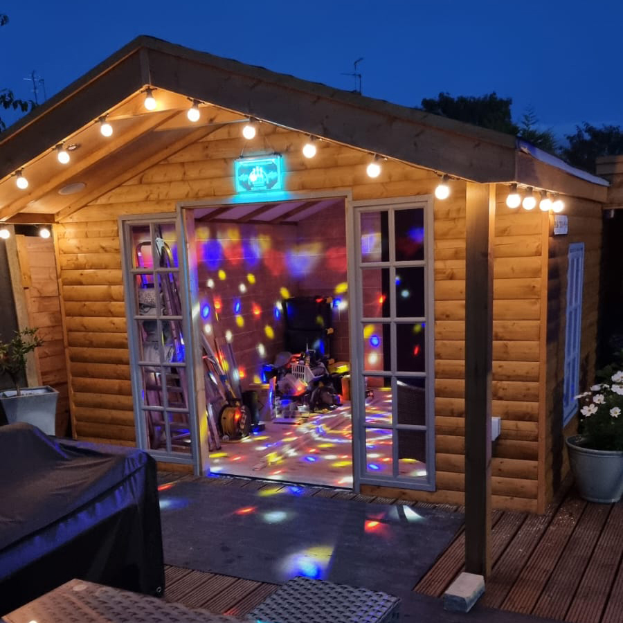 Wooden garden shed at dusk with string lights; colorful disco lights projected inside and on the deck.